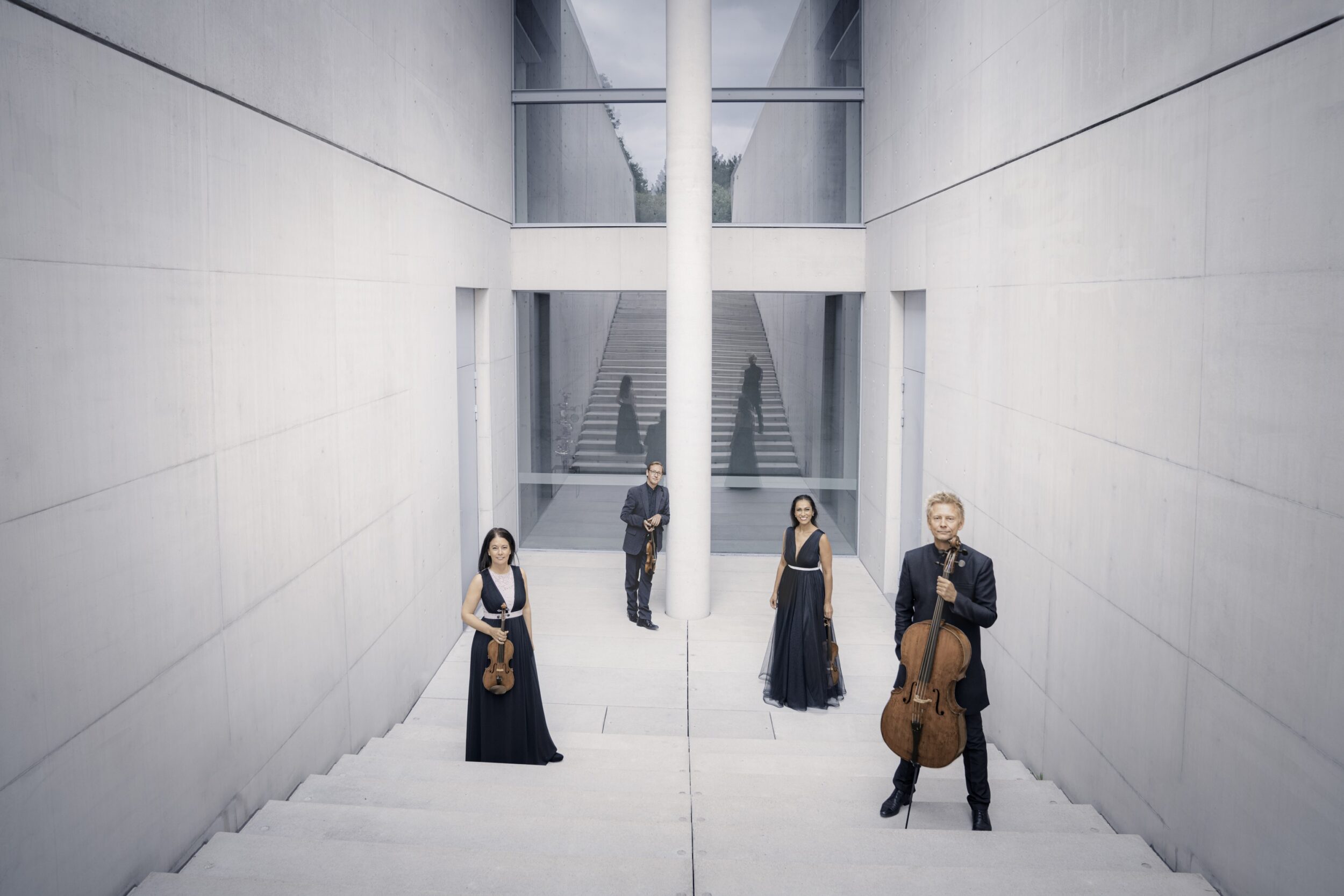 Auf dem Bild sieht man zwei Frauen und zwei Männer. Sie sind das Minguet Quartett. Sie stehen getrennt voneinander auf einer weißen Treppe und halten ihre Musikinstrumente in der Hand. Alle sind in Abendkleidung gekleidet.