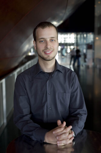 Das Porträt eines Mannes (Ondřej Adámek). Er sitzt an seinem Schreibtisch, die Hände vor sich gefaltet. Er blickt geradeaus und trägt ein schwarzes Hemd. Im Hintergrund ist das Innere eines öffentlichen Gebäudes zu erkennen.